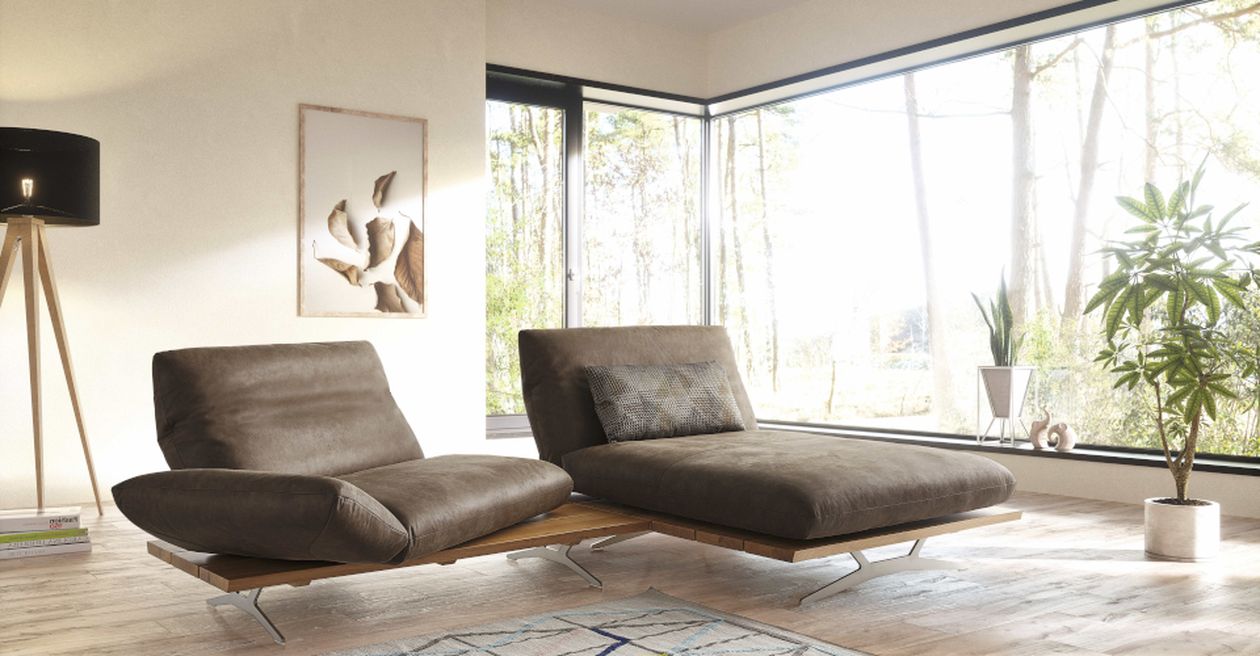 Modernes Wohnzimmer mit zwei braunen Liegesesseln, großen Fenstern mit Blick auf Bäume, einer Stehlampe und einer Zimmerpflanze in der Ecke.