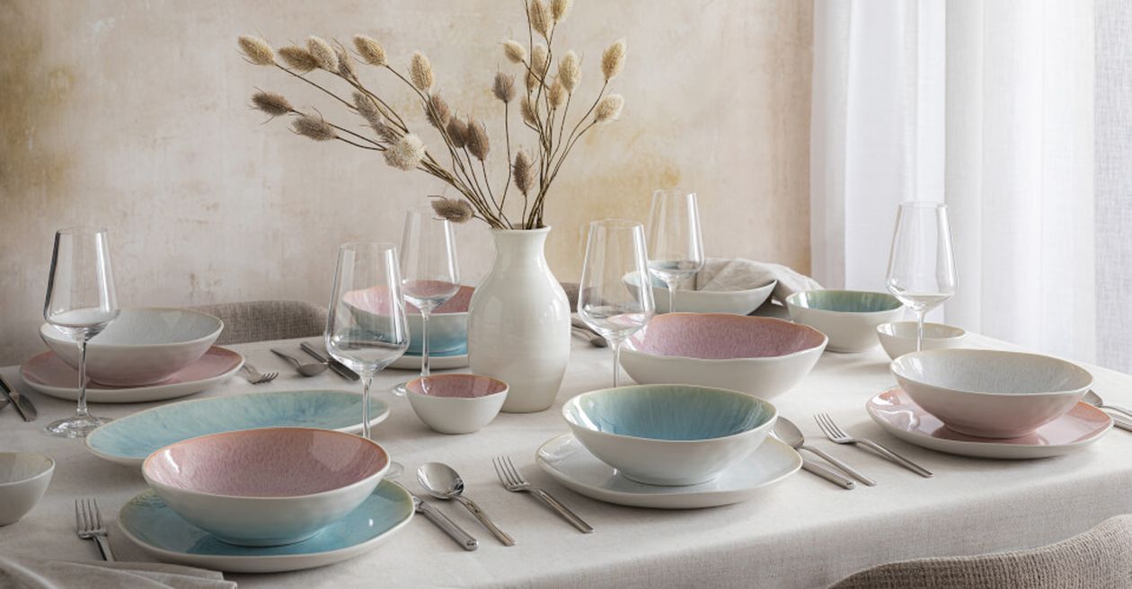 Pastellfarbene Tischdekoration mit Tellern, Gläsern, Besteck und einem Blumenarrangement in einer Vase auf einem gedeckten Tisch.