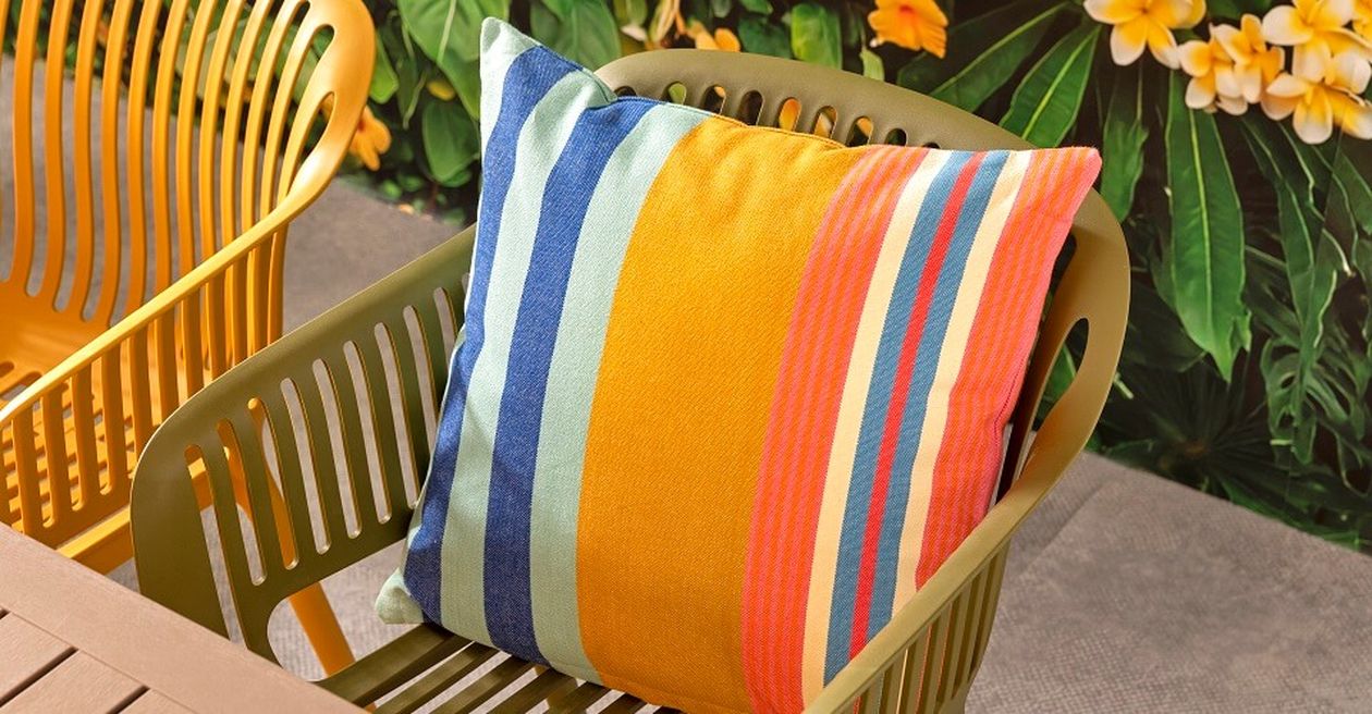 Ein farbenfroh gestreiftes Kissen auf einem grünen Stuhl neben einem Holztisch, vor dem Hintergrund tropischer Blätter und gelber Blüten.