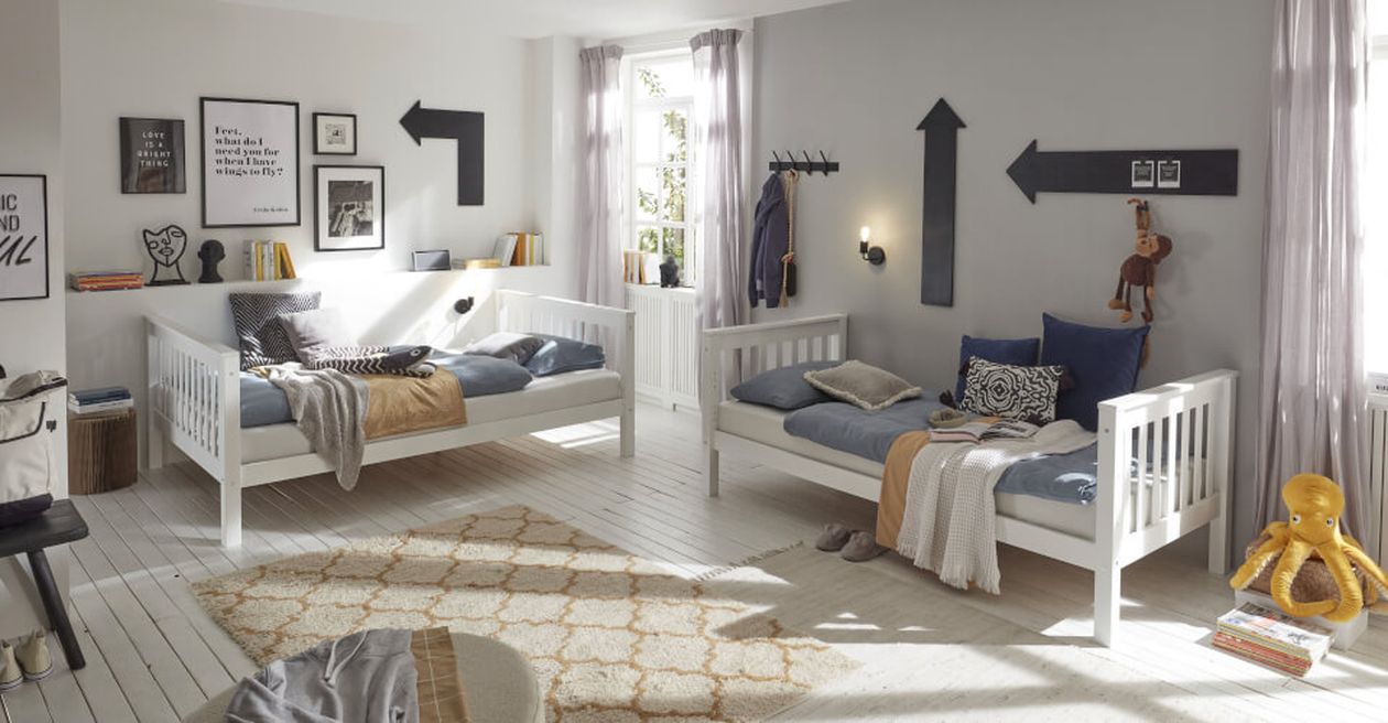 Ein gemütliches Kinderzimmer mit zwei weißen Betten, verspielter Dekoration, Wandbildern und Spielzeug. Licht fällt durch ein Fenster und schafft eine warme Atmosphäre.