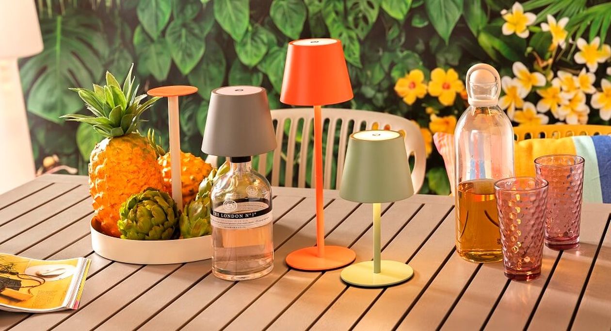 Ein stilvoller Tisch mit farbenfrohen Lampen, einer Obstplatte mit Ananas und Artischocken, einer Glasflasche und zwei Gläsern vor einem floralen Hintergrund.