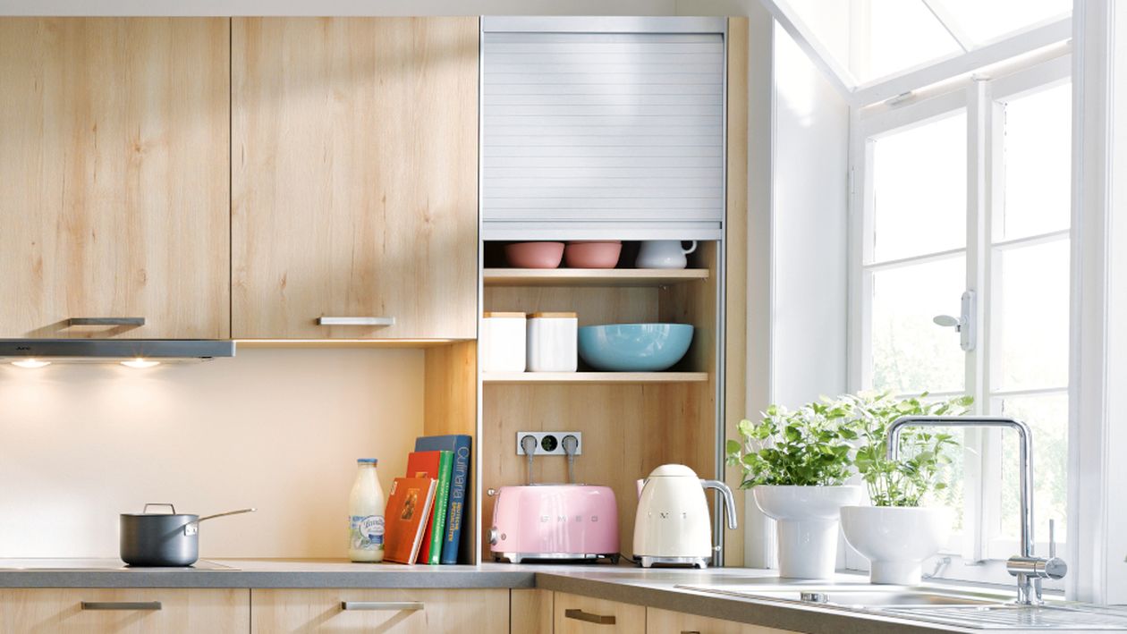 Helle, moderne Küche mit hellen Holzschränken, rosa Toaster auf der Arbeitsplatte und weißem Wasserkocher, blauer Schüssel, Topfkräuter neben der Spüle und sonnendurchflutetem Fenster.