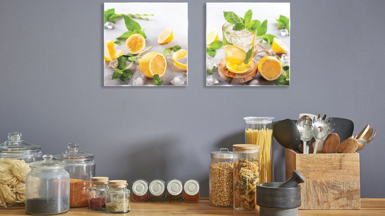 Küchentheke mit Glasbehältern mit Nudeln, Getreide und Gewürzen, einem hölzernen Besteckhalter und zwei Fotos mit Zitronenmotiven an einer grauen Wand.