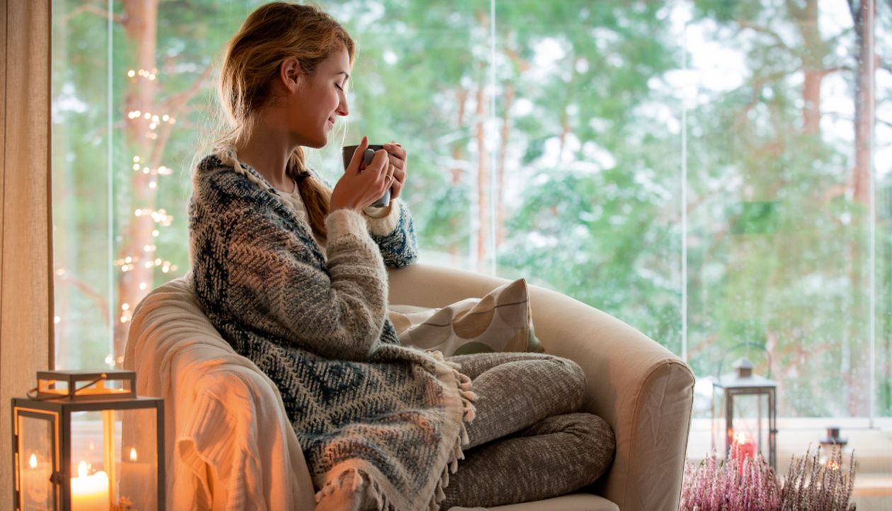 Frau sitzt entspannt in einem Sessel mit Tasse in der Hand, eingekuschelt in eine Decke, vor einem großen Fenster mit Blick auf einen Wald. Im Raum stehen Laternen und Kerzen für eine gemütliche Atmosphäre.