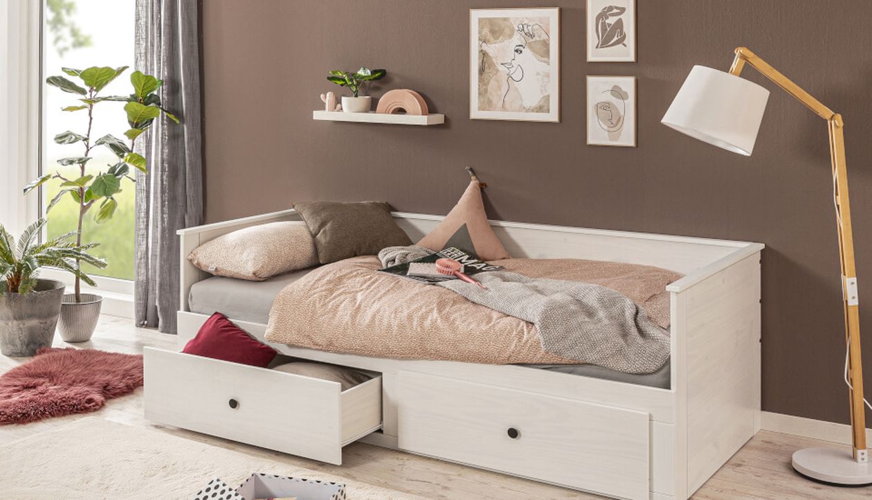 Gemütliches Schlafzimmer mit weißem Tagesbett mit Auszugsschubladen, rosa Bettwäsche, Stehlampe, Wandbildern und Topfpflanzen in Fensternähe.