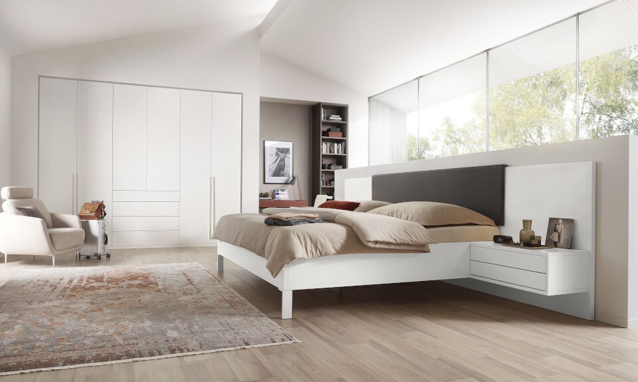 Modernes Schlafzimmer mit großem Bett, beiger Bettwäsche, weißen Möbeln, einem grauen Sessel, einem weichen Teppich und einer Fensterfront mit Blick ins Grüne.