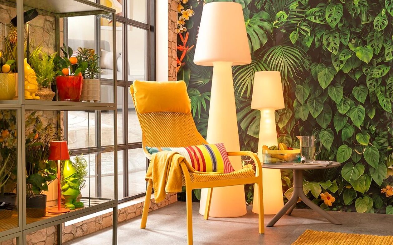 Gemütlicher Outdoor-Bereich mit gelbem Sessel, gestreiftem Kissen, Stehlampen und grüner Blättertapete. Regale mit Pflanzen und Dekorationen stehen an einem großen Fenster.