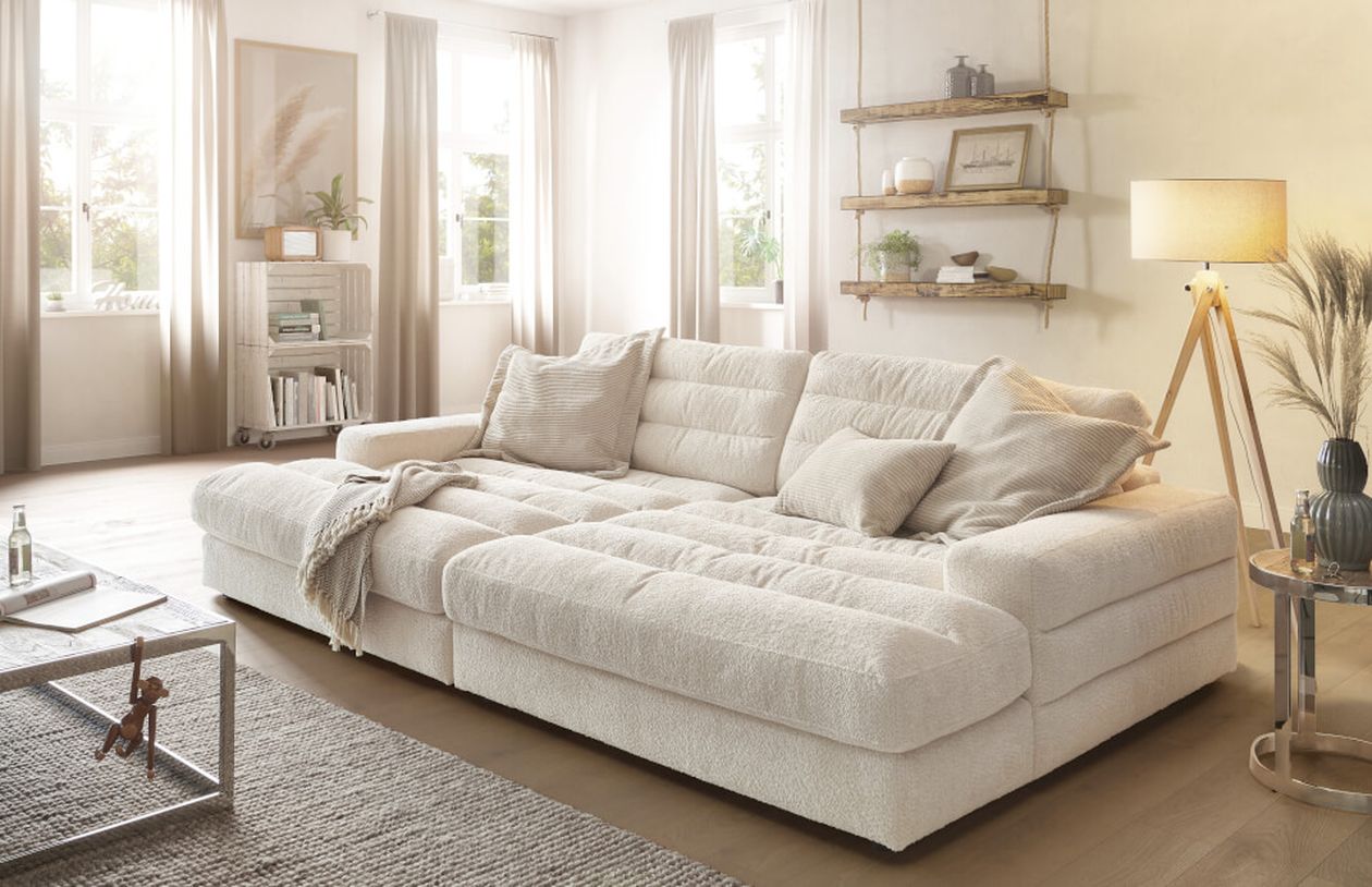 Ein gemütliches Wohnzimmer mit einem großen weißen Ecksofa, beigefarbenen Kissen, einer Stehlampe und schwebenden Regalen. Natürliches Licht strömt durch die Fenster.