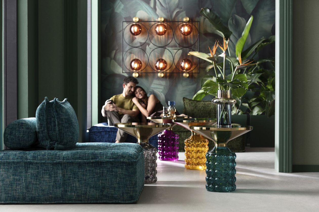Wohnzimmer mit Sofa, Ledersessel und rundem Couchtisch auf einem gemusterten Teppich. Pflanzen, afrikanische Deko-Elemente und eine große Hängeleuchte schaffen eine tropische Atmosphäre.