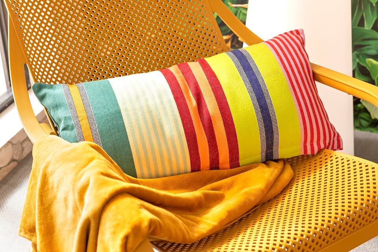 Ein Korbsessel mit einem bunt gestreiften Kissen und einer senfgelben Decke, in der Nähe eines Fensters, im Hintergrund Grün.