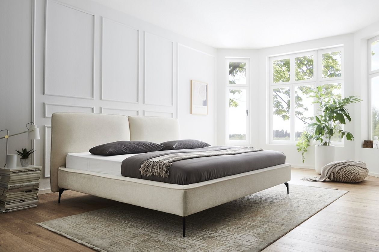 Ein modernes Schlafzimmer mit beigem Bett, grauer Bettwäsche, großen Fenstern, einer Topfpflanze und einem Bücherstapel auf Holzboden.