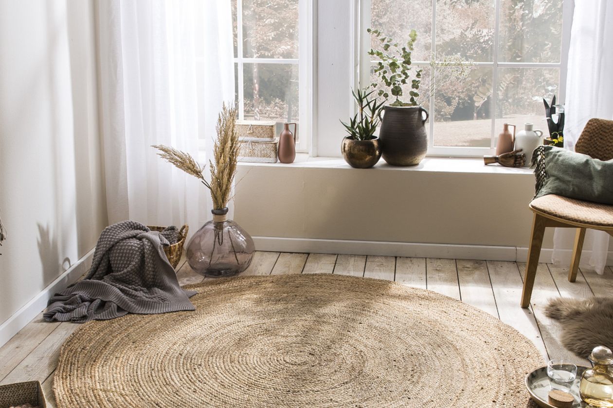 Gemütliche Sitzecke mit Juteteppich, grauer Decke, Pflanzen auf der Fensterbank und Blick auf einen hellen Garten.