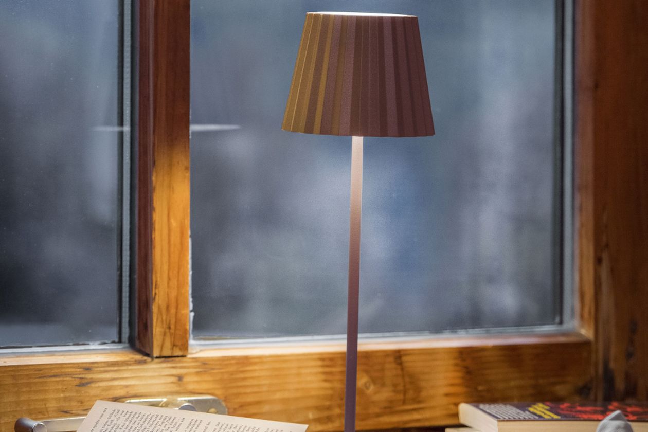 Schlichte Tischlampe mit braunem Lampenschirm auf einem Holztisch vor einem Fenster mit Blick nach draußen.