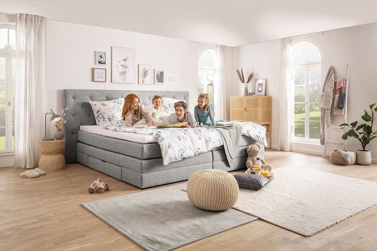 Eine vierköpfige Familie lächelt und sitzt auf einem Bett in einem hellen, gemütlichen Schlafzimmer mit moderner Einrichtung und großen Fenstern.