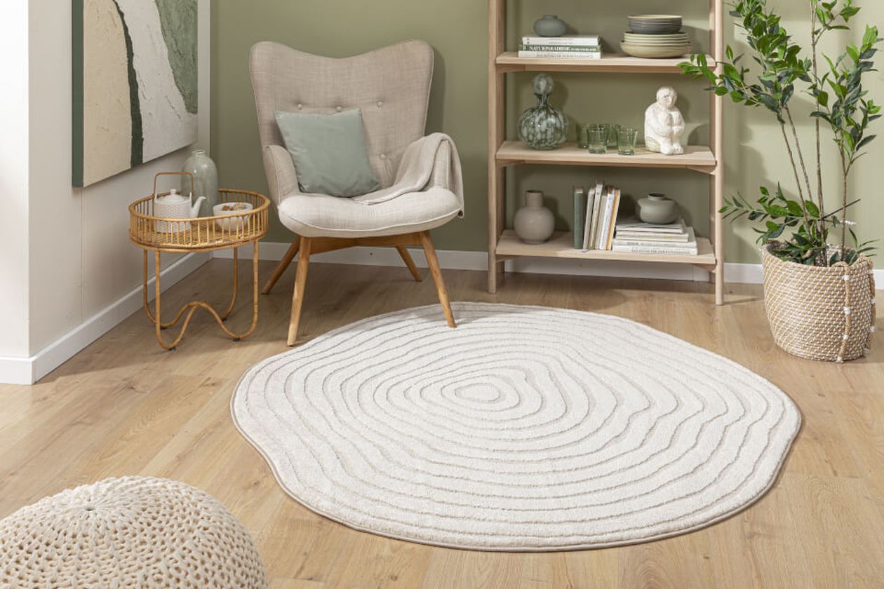 Gemütliches Wohnzimmer mit einem grauen Sessel, einem runden, strukturierten Teppich, Holzregalen mit Dekoration und Topfpflanzen auf hellem Holzboden.