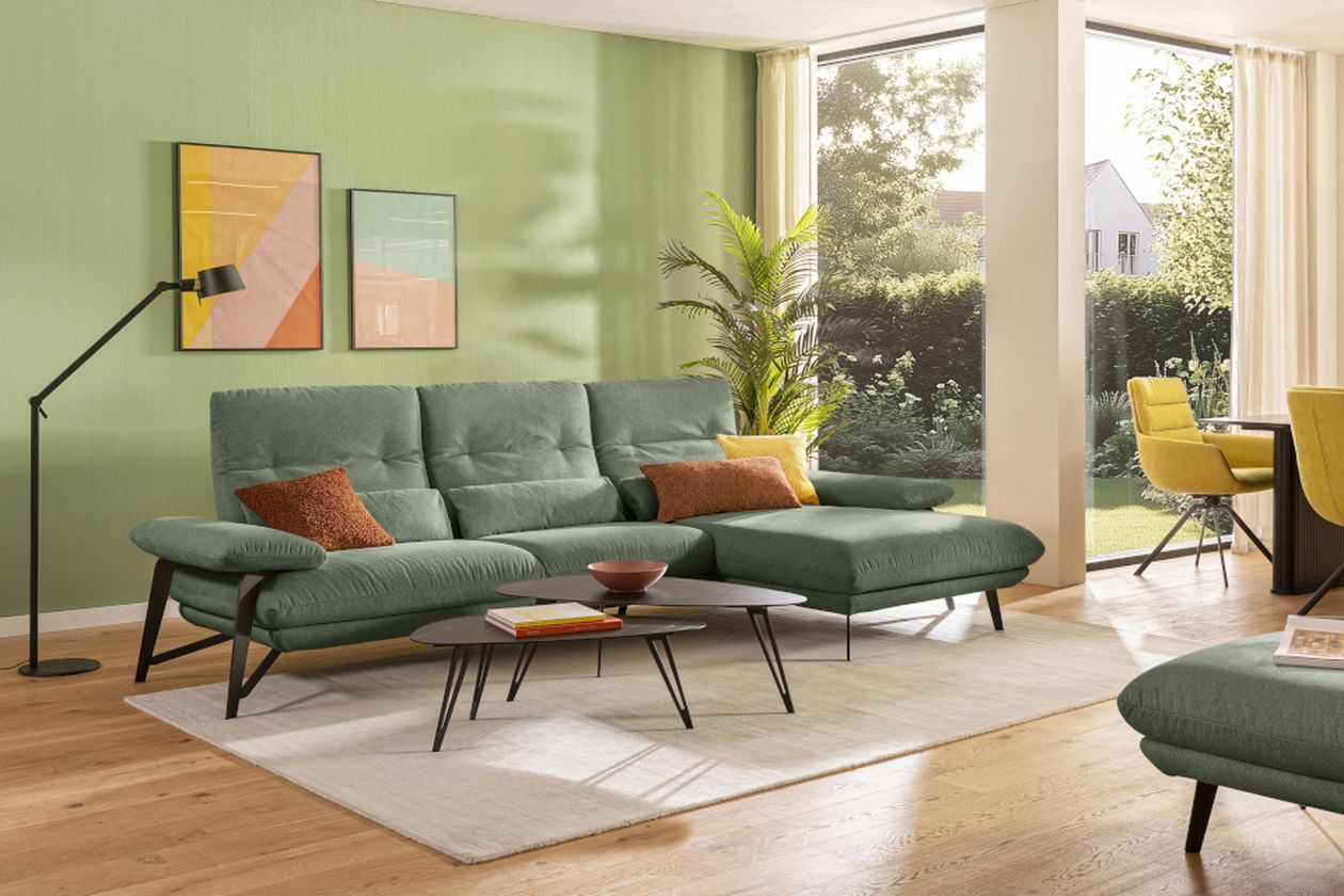 Helles Wohnzimmer in Pastellgrün: große Eckcouch mit farbigen Kissen, zwei Couchtische und moderne Stehlampe. Große Fenster mit Blick ins Grüne.