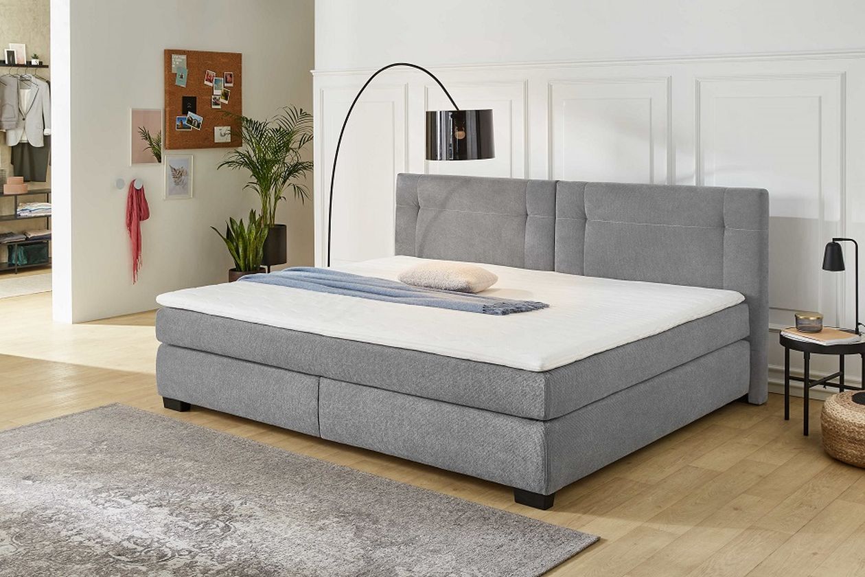 Ein modernes Schlafzimmer mit einem großen grauen Polsterbett, einer Stehlampe und einem Teppich auf Holzboden. Im Hintergrund sind eine Pflanze und eine Pinnwand zu sehen.