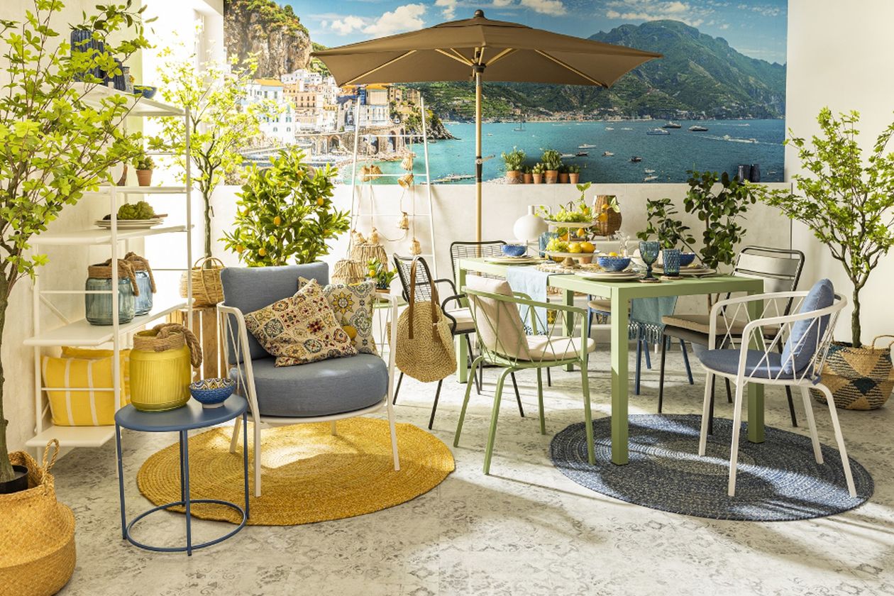Sonnige Terrasse am Meer mit Sonnenschirm, gedecktem Esstisch, verschiedenen Stühlen und Liegestuhl mit Kissen, Topfpflanzen und Meerblick im Hintergrund.