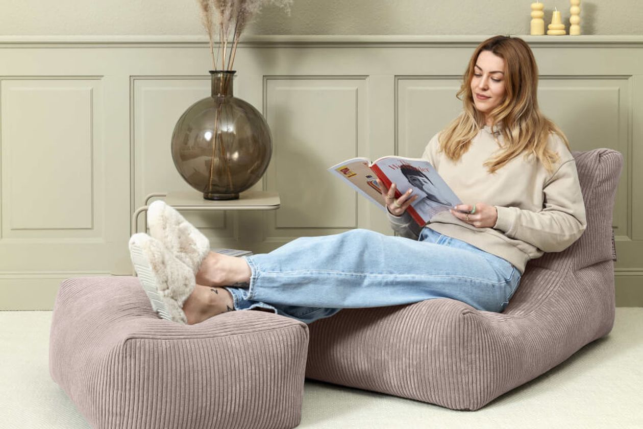 Eine Frau entspannt auf einer weichen, taupefarbenen Liege und liest eine Zeitschrift. Sie trägt einen beigen Pullover, Jeans und flauschige Hausschuhe. Die Einrichtung ist minimalistisch.