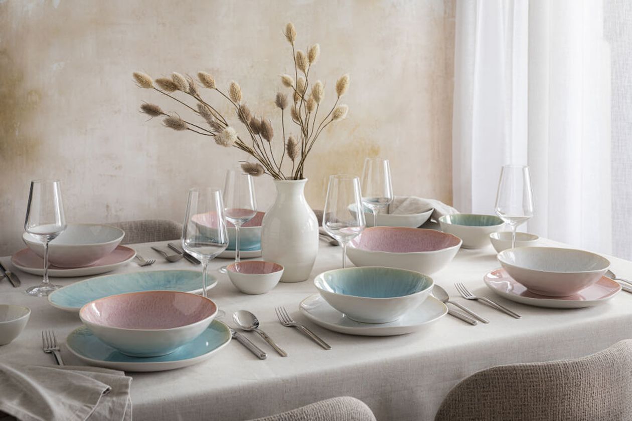 Pastellfarbene Tischdekoration mit Tellern, Gläsern, Besteck und einem Blumenarrangement in einer Vase auf einem gedeckten Tisch.