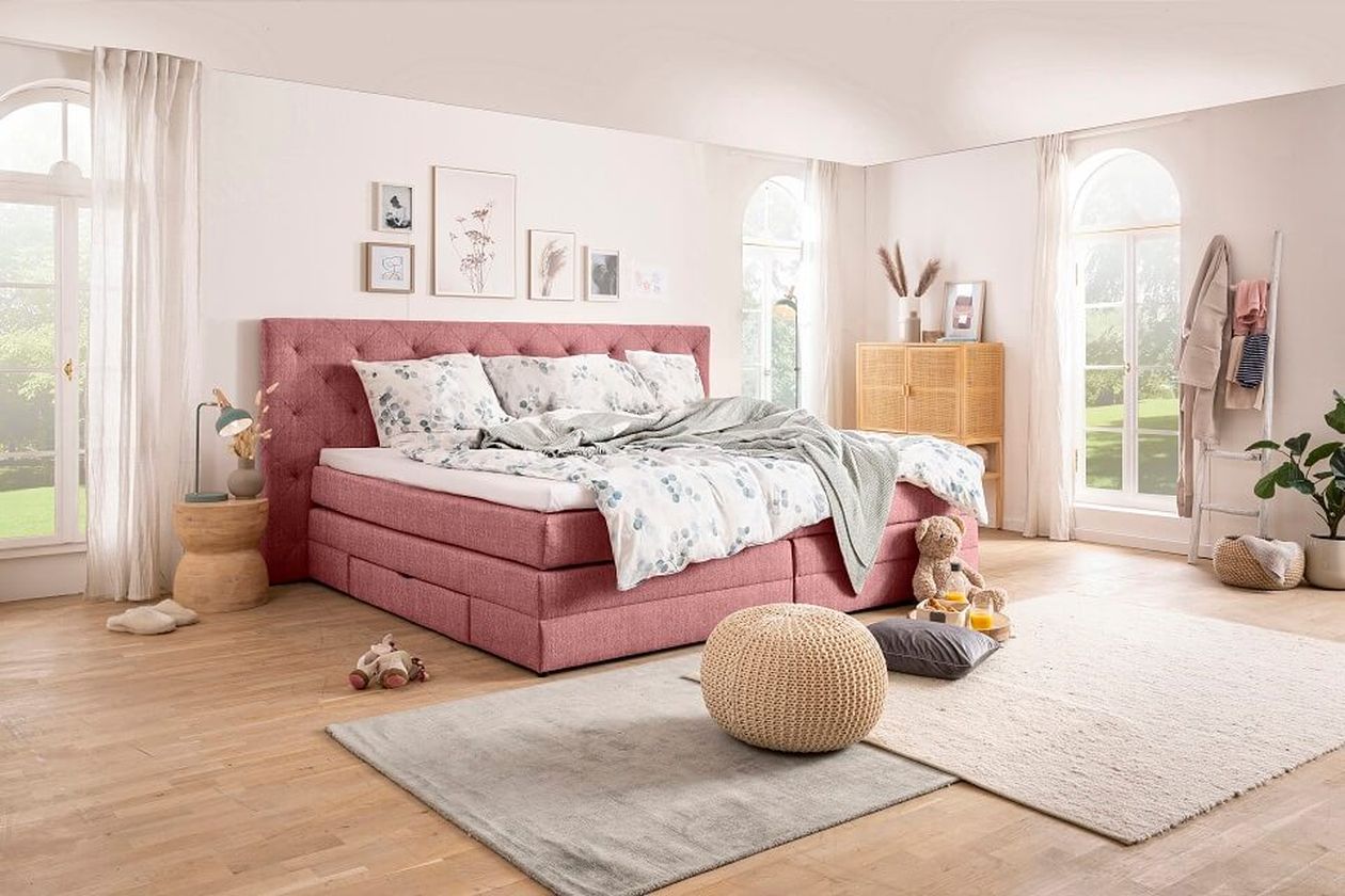 Helles Schlafzimmer mit rosa Polsterbett, heller Bettwäsche und dekorativen Kissen. Holzfußboden, große Fenster und minimalistische Deko.