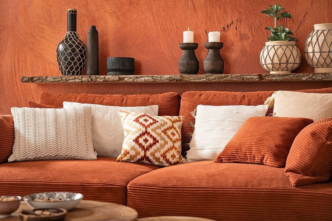 Gemütliches Wohnzimmer mit Terrakotta-Sofa, verschiedenen Kissen, rustikalem Regal mit Vasen und Kerzen sowie einer warmfarbigen, strukturierten Wand.