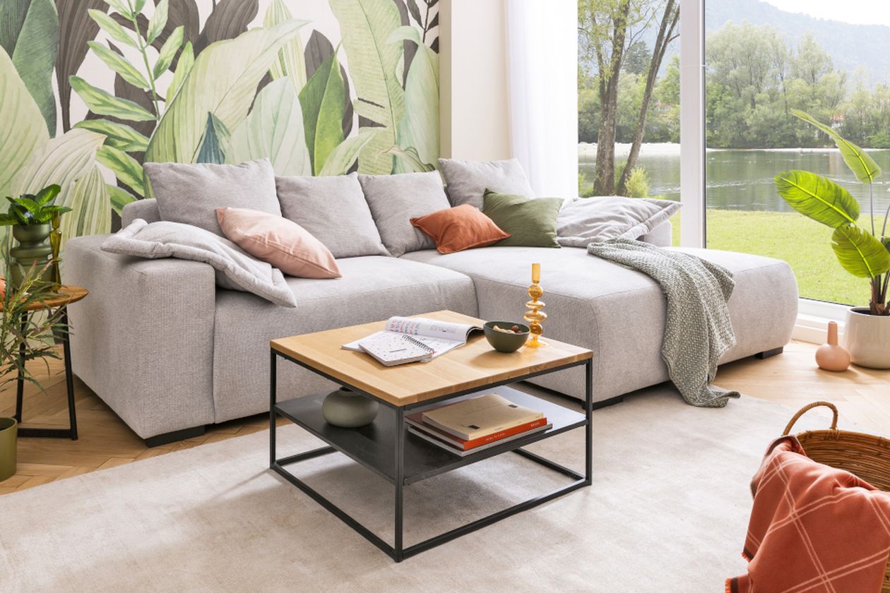 Helles Wohnzimmer mit grauer Eckcouch, Holztisch und großen Fenstern mit Blick ins Grüne. Bananenblätter-Tapete, Pflanzen und bunte Kissen schaffen eine natürliche Atmosphäre.