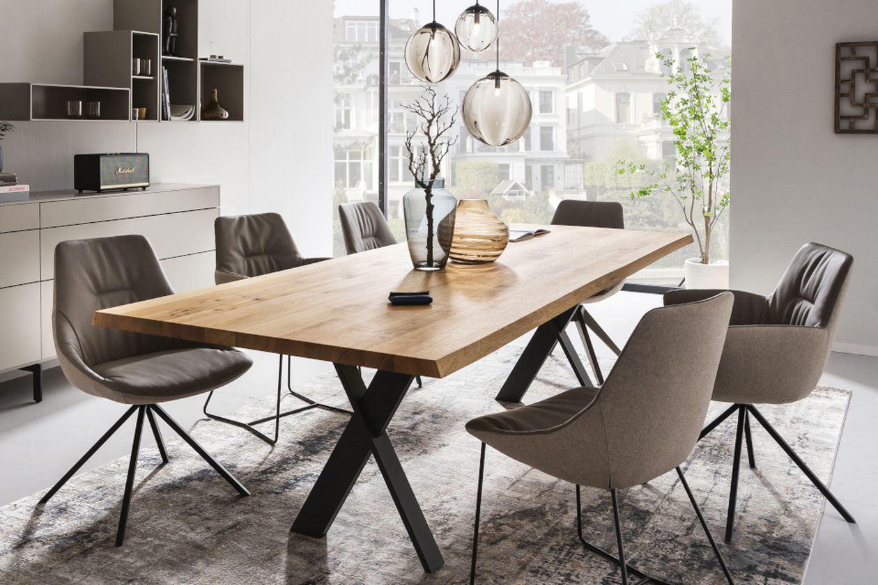 Modernes Esszimmer mit langem Holztisch auf schwarzen X-Beinen, sechs grau gepolsterten Stühlen, Glaspendelleuchten und Vasen an einem großen Fenster.