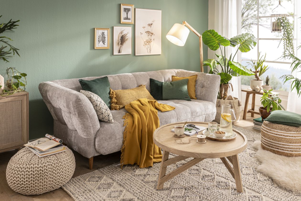 Gemütliches Wohnzimmer in Mintgrün: graue Couch mit gelben und grünen Kissen, Couchtisch mit Tee-Arrangement. Pflanzen, Bilder und gemusterter Teppich runden den Raum ab.