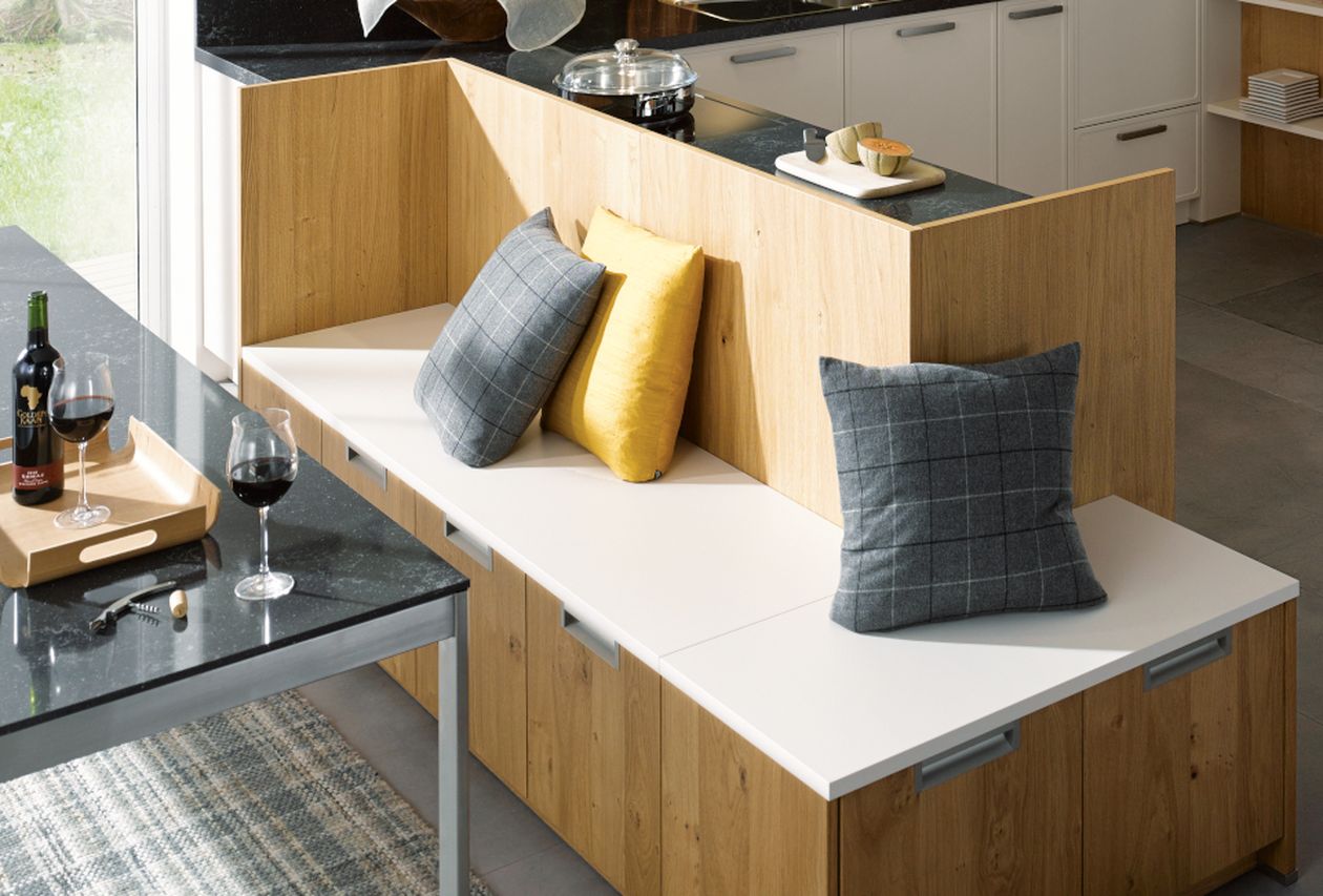 Moderne Küchenecke mit eingebauter Holzbank, weißem Sitz, grauen und gelben Kissen, daneben ein Tisch mit Weinflasche.