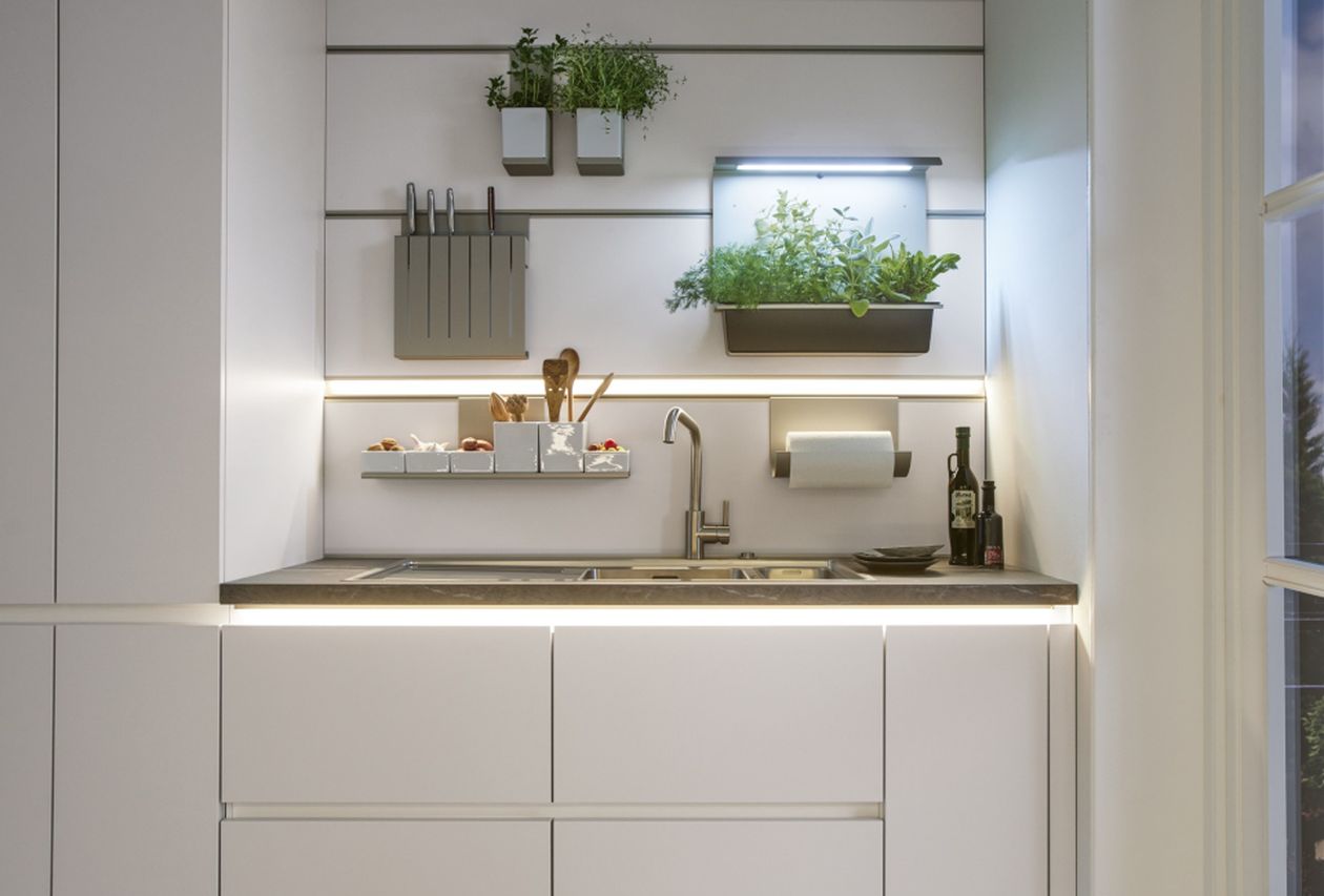 Minimalistische weiße Küchenspüle mit Edelstahlarmatur, Messerblock, Wandkräuterkästen, Unterbaubeleuchtung und Flaschen auf der Arbeitsplatte.