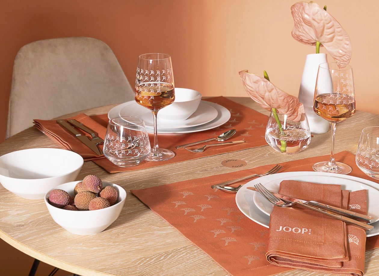 Elegant gedeckter Tisch mit orangefarbenen Platzsets, Gläsern und weißem Geschirr. Rosa Blumen in Vasen und eine Schale mit Litschis runden das Dekor ab.