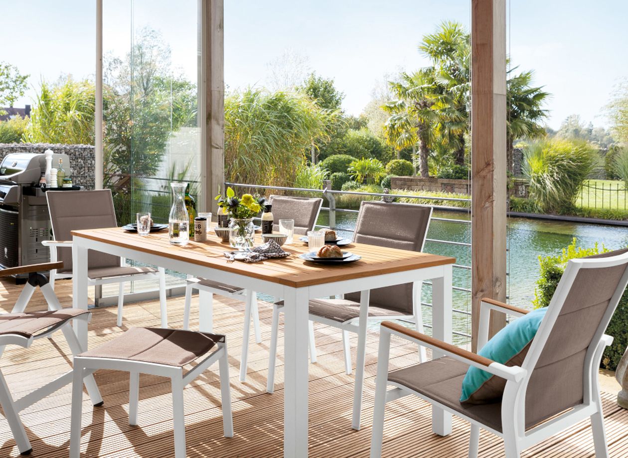 Terrasse mit weißem Esstisch, braunen Stühlen und Blick auf einen Garten mit Teich, Palmen und Grillbereich.