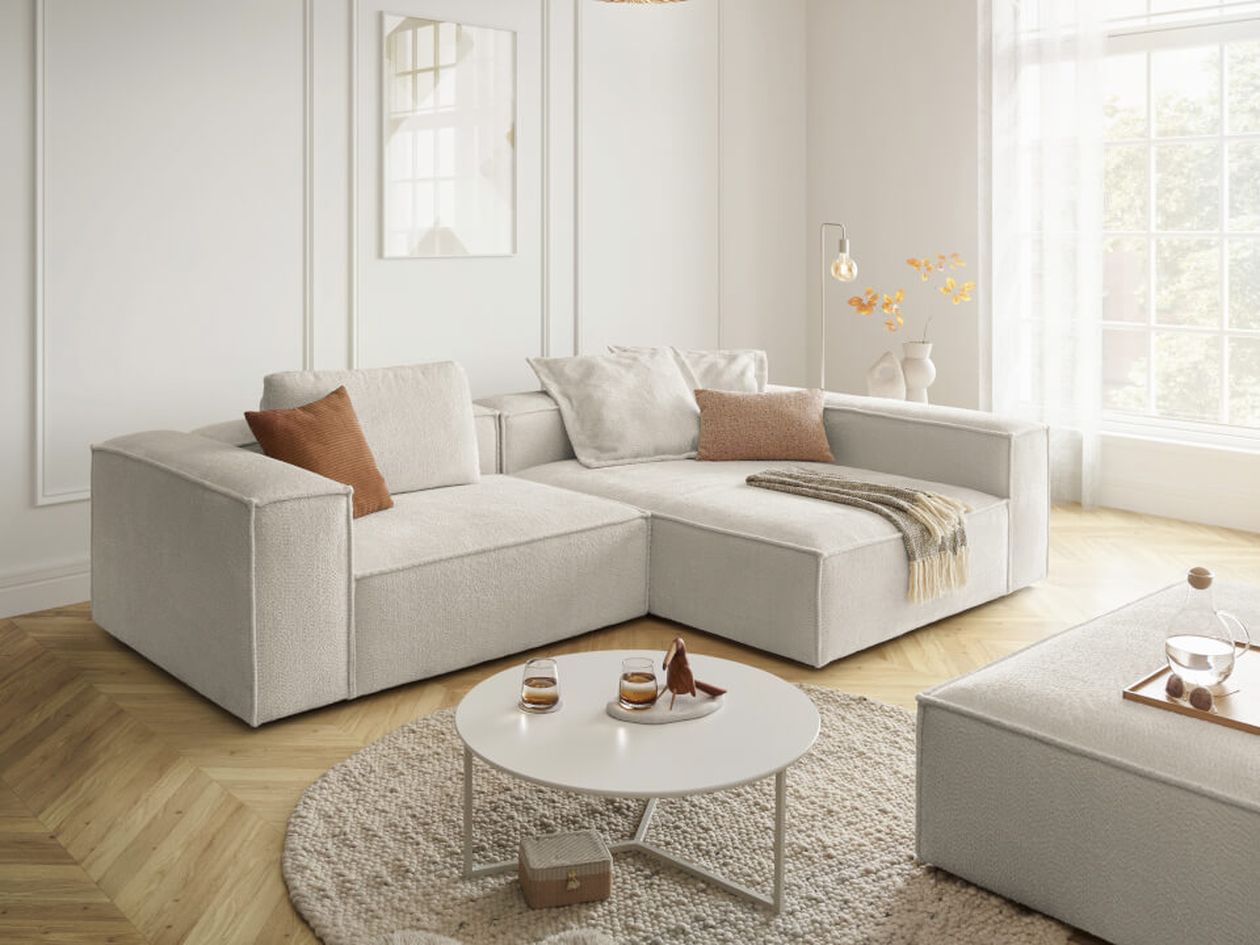 Modernes Wohnzimmer mit hellgrauem Ecksofa, braunen Kissen, einem runden weißen Couchtisch und sanftem natürlichem Licht durch ein großes Fenster.