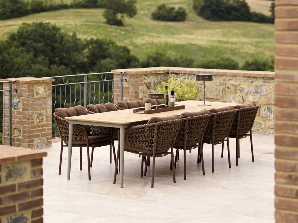 Langer hölzerner Esstisch für den Außenbereich mit acht geflochtenen Stühlen, Weinflasche und Gläsern auf einer Steinterrasse mit Blick auf die Landschaft.