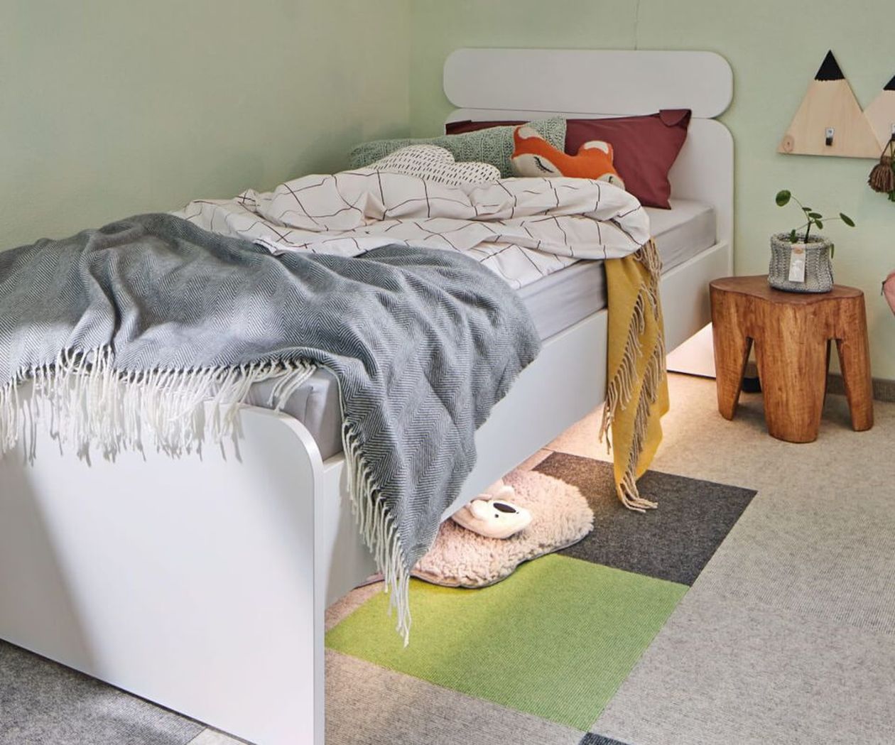 Gemütliches Schlafzimmer mit weißem Bett, farbenfroher Bettwäsche, grauer Tagesdecke, Holzhocker, Topfpflanze und geometrisch gemustertem Teppich auf dem Boden.