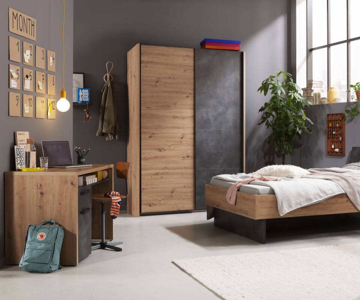 Modernes Schlafzimmer mit Holzmöbeln, darunter Bett, Schreibtisch und Kleiderschrank. Laptop und Rucksack stehen auf dem Schreibtisch. Großes Fenster und Wandbilder.