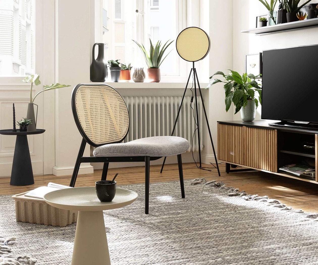 Modern living room with a woven chModernes Wohnzimmer mit einem Korbsessel, einem runden Tisch, Pflanzen, einer Stehlampe, einem Fernseher und einem strukturierten Teppich auf Holzboden.air, round table, plants, floor lamp, TV, and a textured rug on wooden flooring.