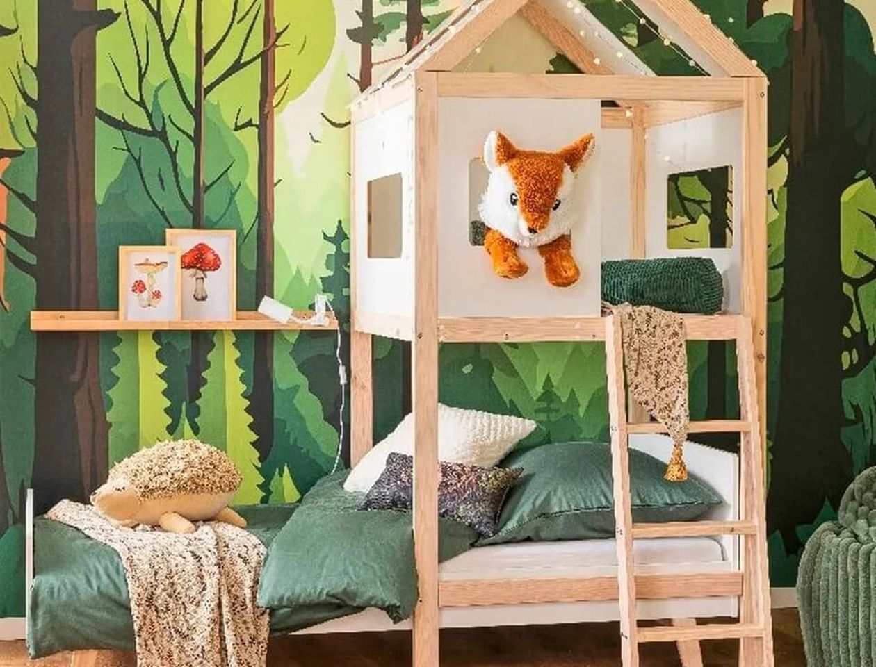 Holzfarbenes Kinderhausbett mit Leiter und Fuchs-Kuscheltier am Fenster, vor einer grünen Waldtapete mit Lichterkette dekoriert, grüne Bettwäsche und Kissen.