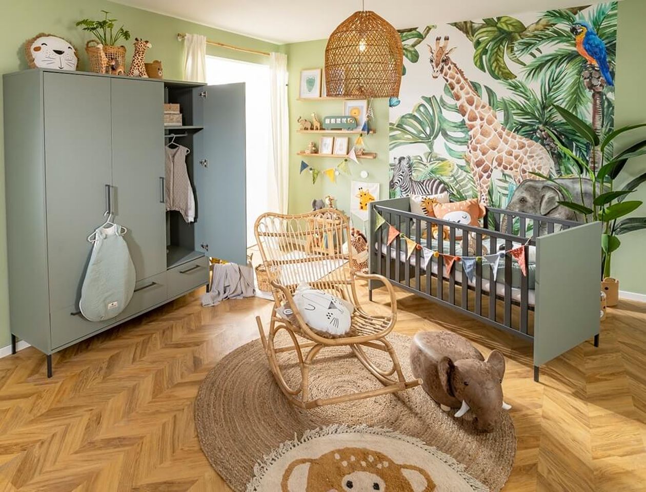 Babyzimmer im Safari-Stil mit grünem Möbelset aus Kleiderschrank und Babybett, Rattanstuhl und Hängelampe auf Fischgrät-Parkett; bunte Dschungel-Fototapete mit Giraffe, Zebra und Elefant, Plüsch-Elefant und Löwen-Kuscheltier als Deko.