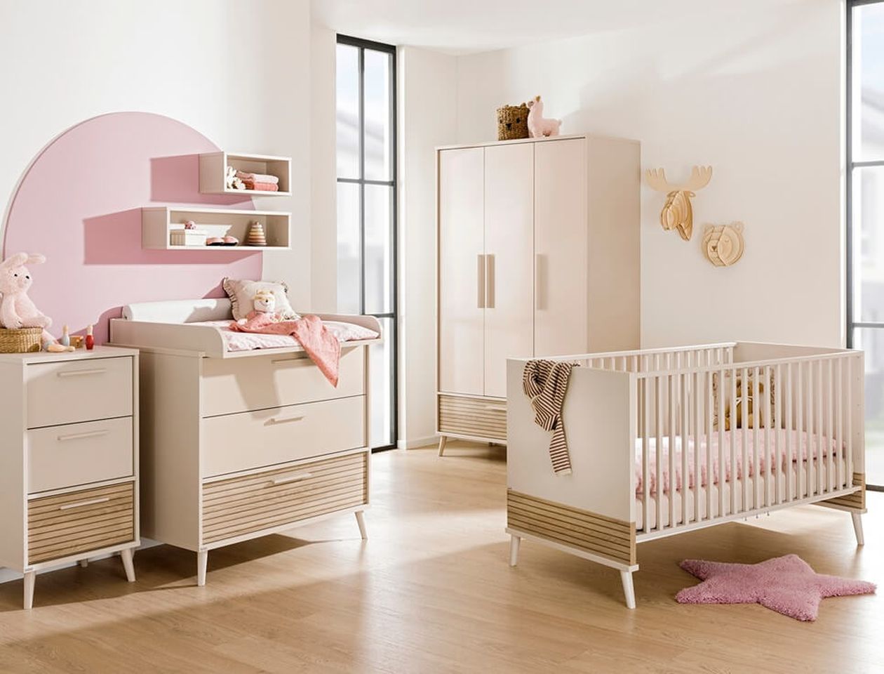 Helles Babyzimmer mit cremefarbenem Möbelset in Weiß-Natur: Wickelkommode mit Aufsatz und Wandregal vor einer rosa Kreisfläche an der Wand, dazu passender Kleiderschrank und Babybett mit Holzakzenten; rosa Stern-Kissen auf dem Boden, Holztier-Wanddeko.