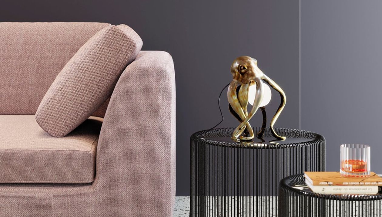 Dekorative Tischleuchte in Form eines goldenen Oktopus, der eine Glaskugel hält, auf einem schwarzen Draht-Beistelltisch neben einem rosafarbenen Sofa.