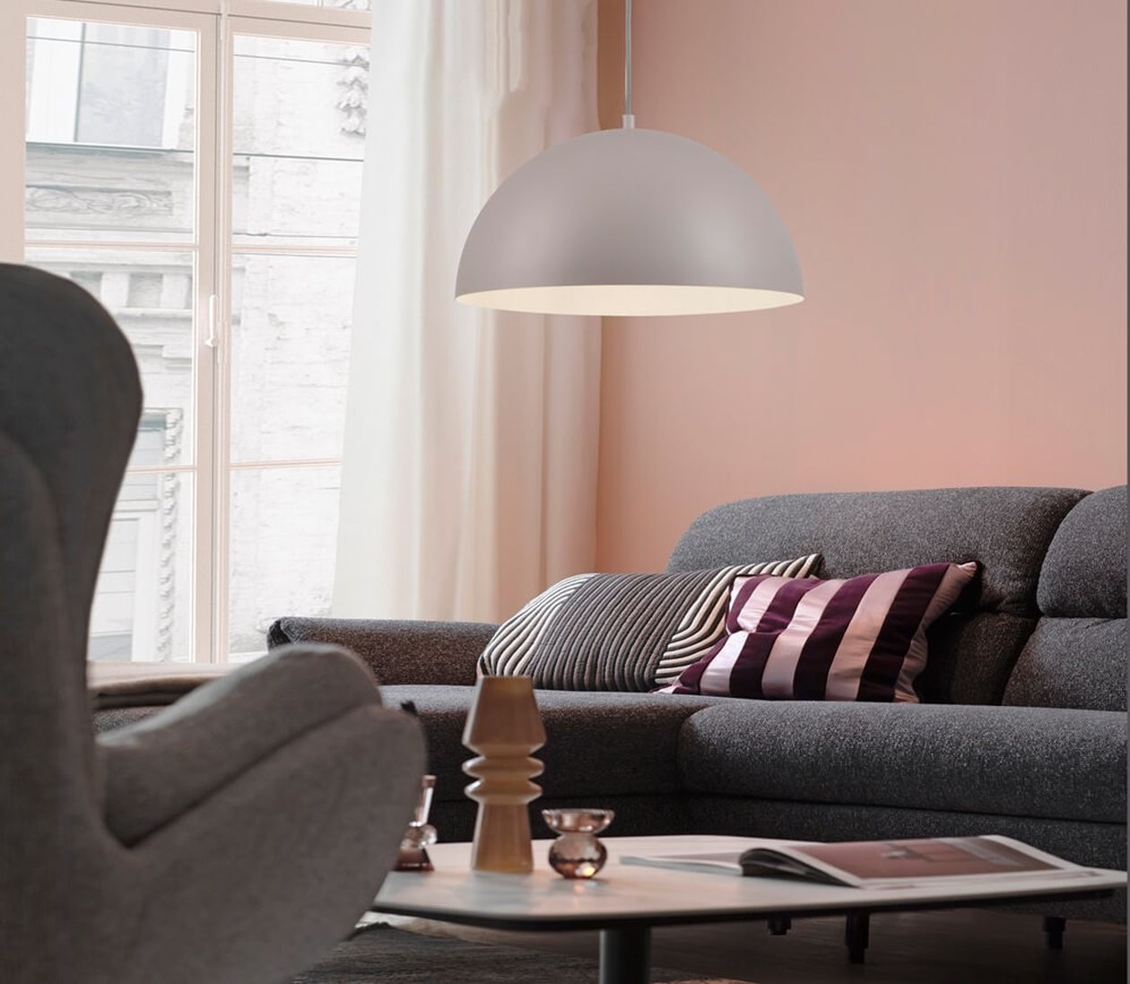 Weiße halbkugelförmige Pendelleuchte aus Metall über einem grauen Sofa mit gestreiften Kissen in einem Wohnzimmer mit rosafarbener Wand.