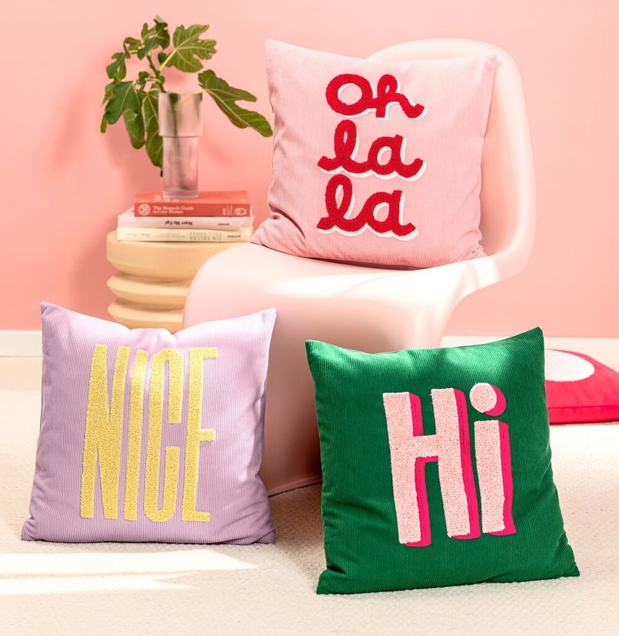 Drei bunte Dekokissen aus Cord mit Schriftzügen: ein rosafarbenes mit „Oh la la", ein fliederfarbenes mit „NICE" und ein grünes mit „Hi", arrangiert auf einem rosa Stuhl.