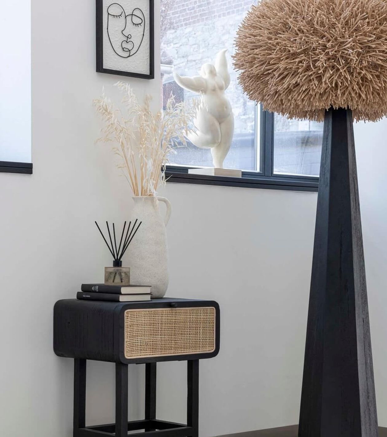 Modernes Interieur mit einem schwarzen Beistelltisch, einer dekorativen Vase, gerahmter Strichzeichnung, einer großen strukturierten Skulptur und einem Fenster mit einer weißen Skulptur davor.