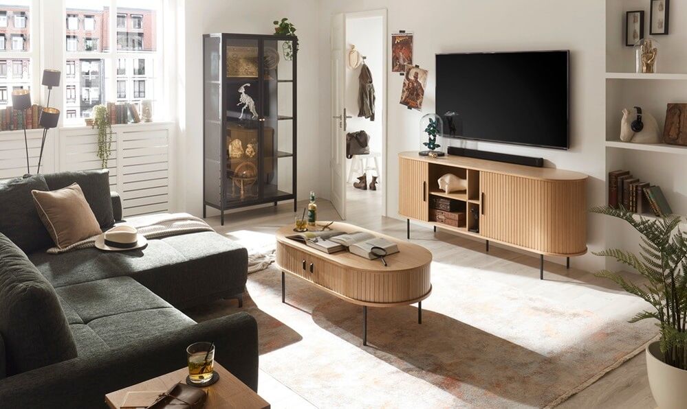 Beigefarbenes TV-Lowboard mit gerillter Front und abgerundeten Seiten in einem Wohnzimmer mit dunkelgrünem Sofa, Vitrine und Fernseher.
