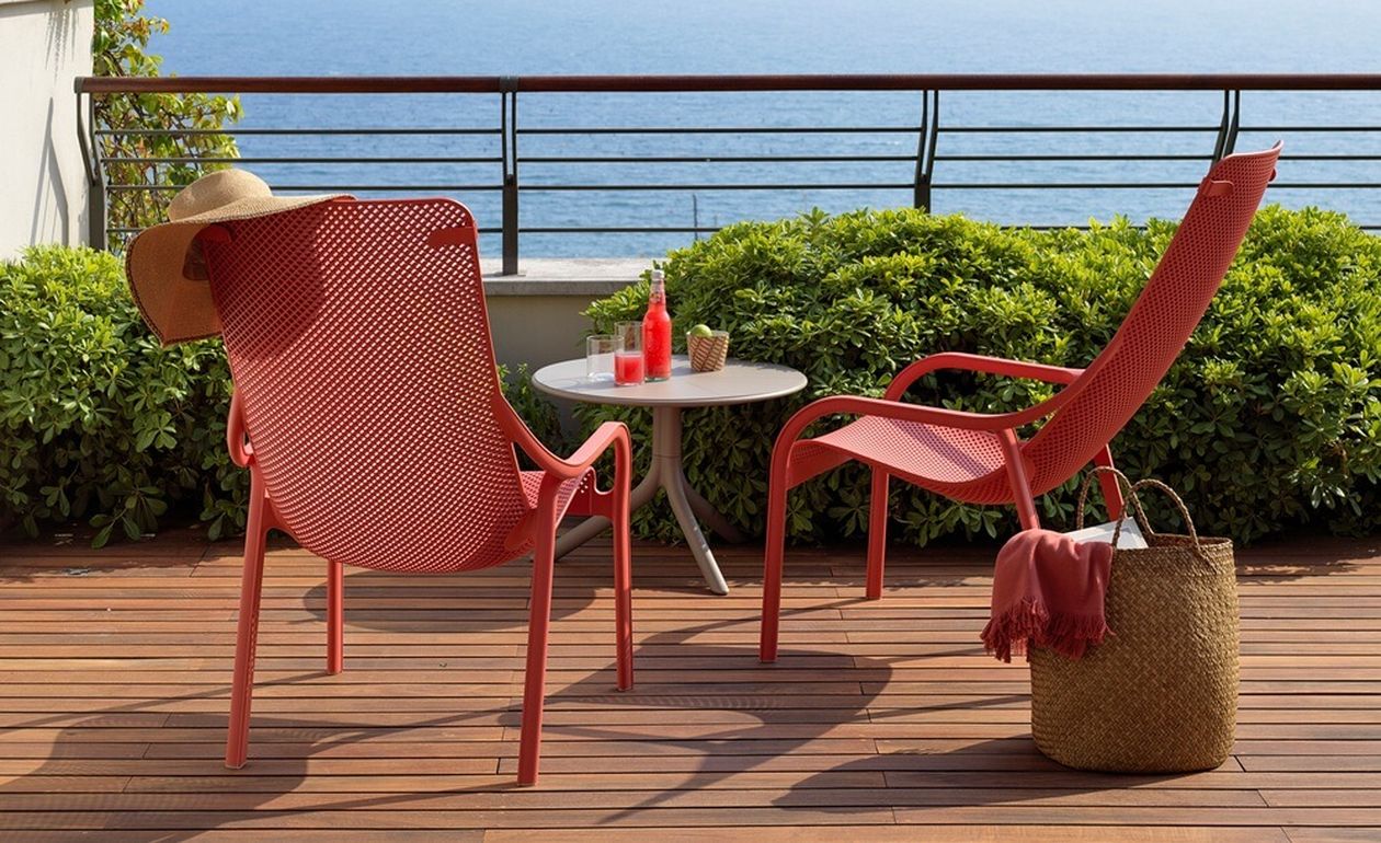 Zwei rote Liegestühle auf einer Holzterrasse mit Blick auf das Meer, mit Getränken auf einem kleinen Tisch, einem Strohhut und einer gewebten Tasche in der Nähe.