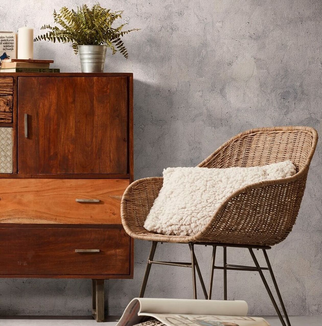 Rattanstuhl mit Metallbeinen und weißem Fellkissen neben einem dunklen Holz-Highboard. Davor ein geflochtener Pouf und eine aufgeschlagene Zeitschrift.