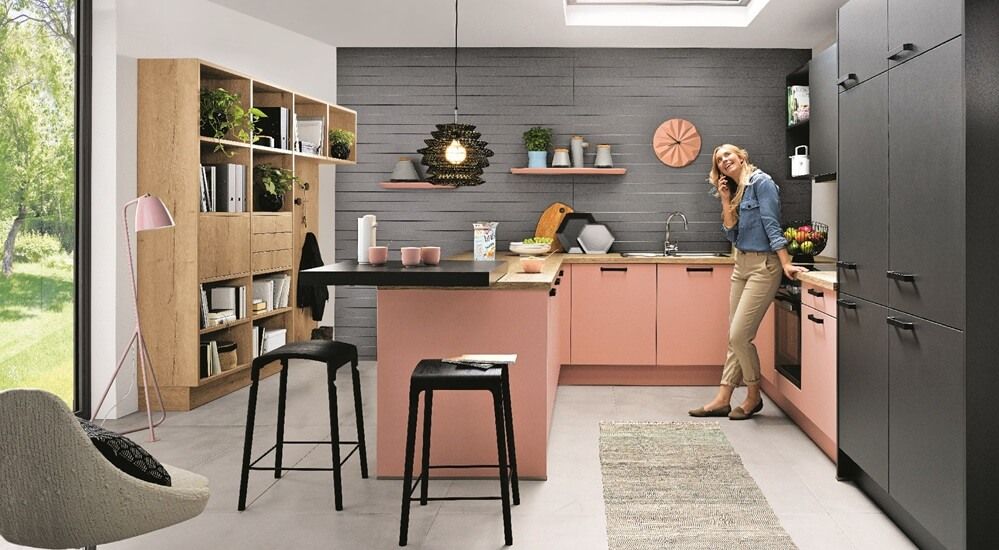 Moderne Küche mit rosa und schwarzen Schränken, einer zentralen Insel mit Hockern und einer Frau, die telefoniert. Großes Fenster mit Blick auf das Grün draußen.