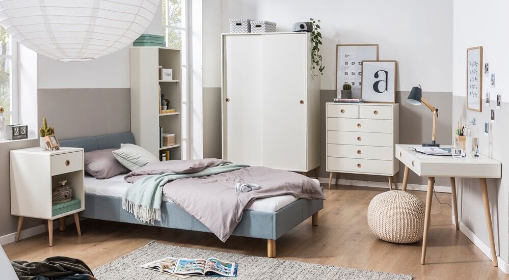 Modernes Schlafzimmer mit Bett, Nachttisch, Kleiderschrank und Schreibtisch. Zur Einrichtung gehören gerahmte Kunst, Bücher und ein Pouf auf Holzboden. Neutrale Töne.